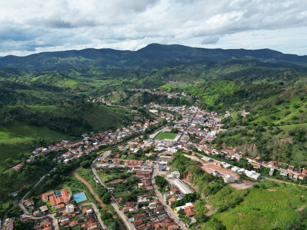 VISTA A&Eacute;REA DE SANTA MARIA DE ITABIRA, cercada por montanhas verdejantes e cortada por um rio sinuoso, destacando a harmonia entre a cidade e a natureza.